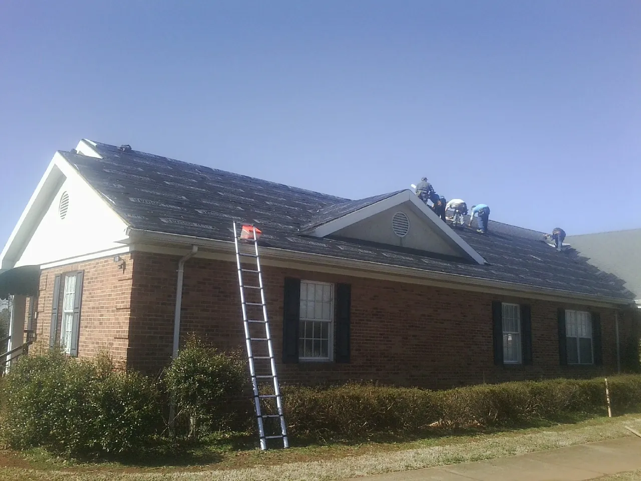 Crew tearing shingles off a brick ranch roof with a ladder set — Metal Roof Repair work by our crew in Prosperity