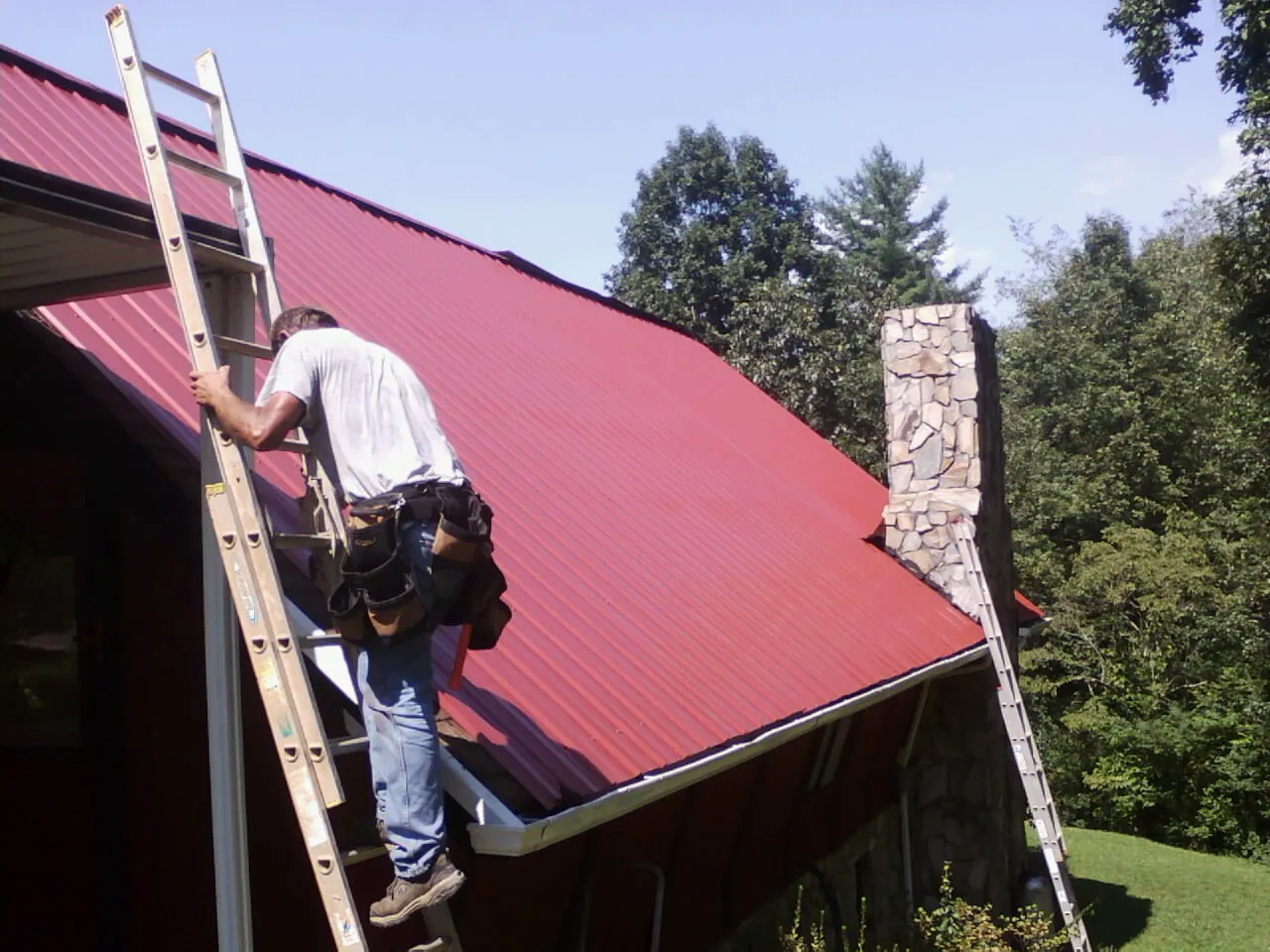 Roofer on a ladder beside a red metal roof and stone chimney — Soffit & Fascia Repair by Roofing Brothers in Prosperity